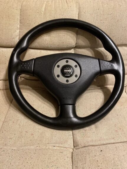 Genuine OEM MOMO steering wheel designed for Mitsubishi Lancer Evolution models (specifically generations Evo IV, V, VI, and Tommi Makinen Edition / CP9A/CN9A platforms), featuring a black leather finish and a central horn pad with the MOMO logo insert.