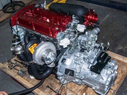 Fully built, high-performance Mitsubishi Lancer Evolution IX engine and manual transmission assembly featuring a red-finished valve cover, a custom exhaust manifold with a large turbocharger, and a billet aluminum transmission mount.