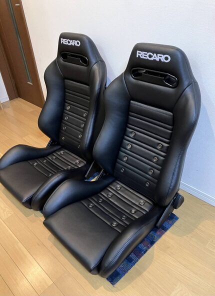 Brand New Pair of Recaro SR3 reclining bucket seats upholstered in black synthetic leather with distinctive horizontal ribbed cushioning and ventilation eyelet details.